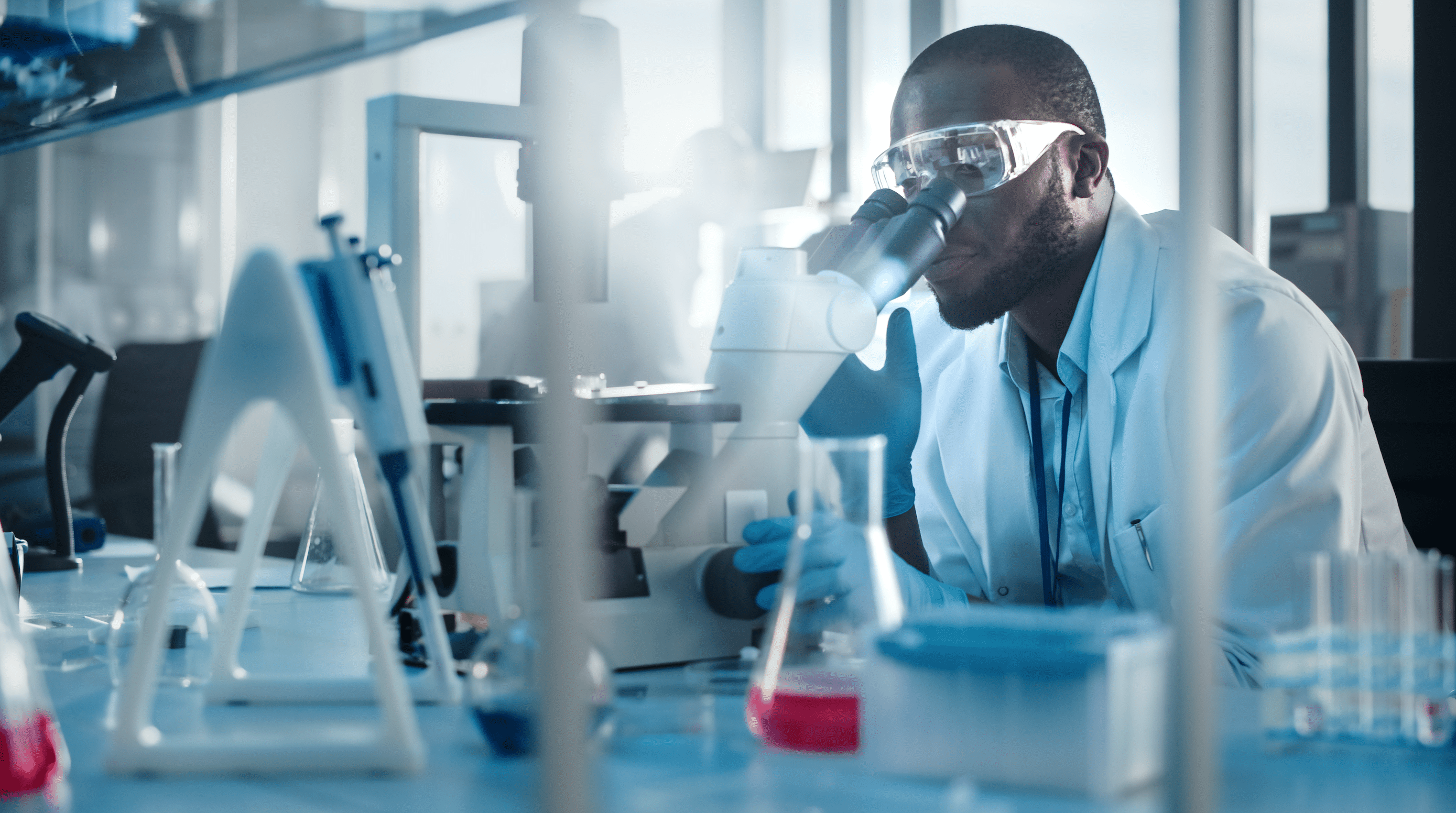 Medical Development Laboratory: Black Male Scientist Looking Under Microscope, Inspecting Petri Dish. Professionals Working in Advanced Scientific Lab doing Medicine, Vaccine, Biotechnology Research